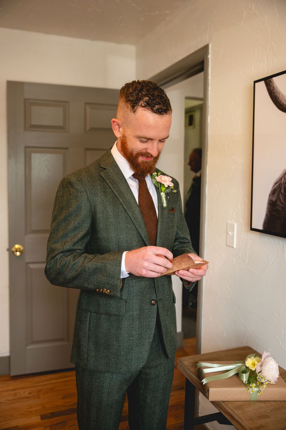 A groom in a green suit and brown tie checks his phone while standing in a room with wooden floors and white walls.
