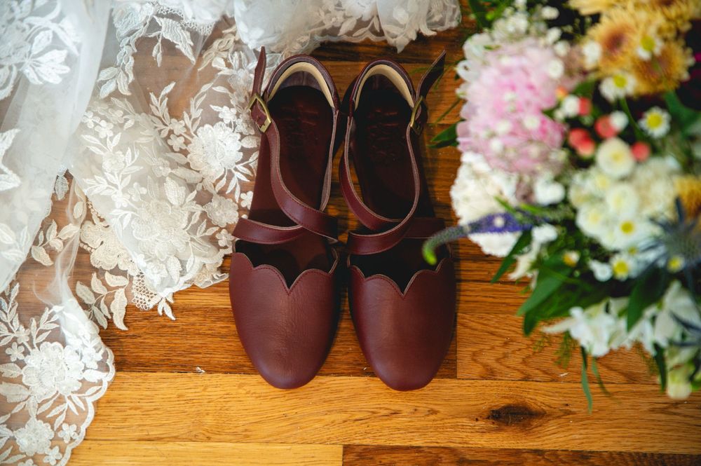Burgundy leather flats placed on wooden floor near white lace and colorful floral decorations.