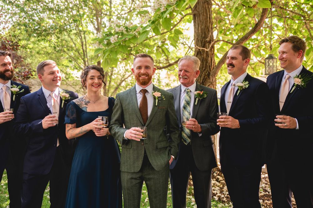 A group of people wearing formal attire laugh together while holding drinks in an outdoor wooded setting.