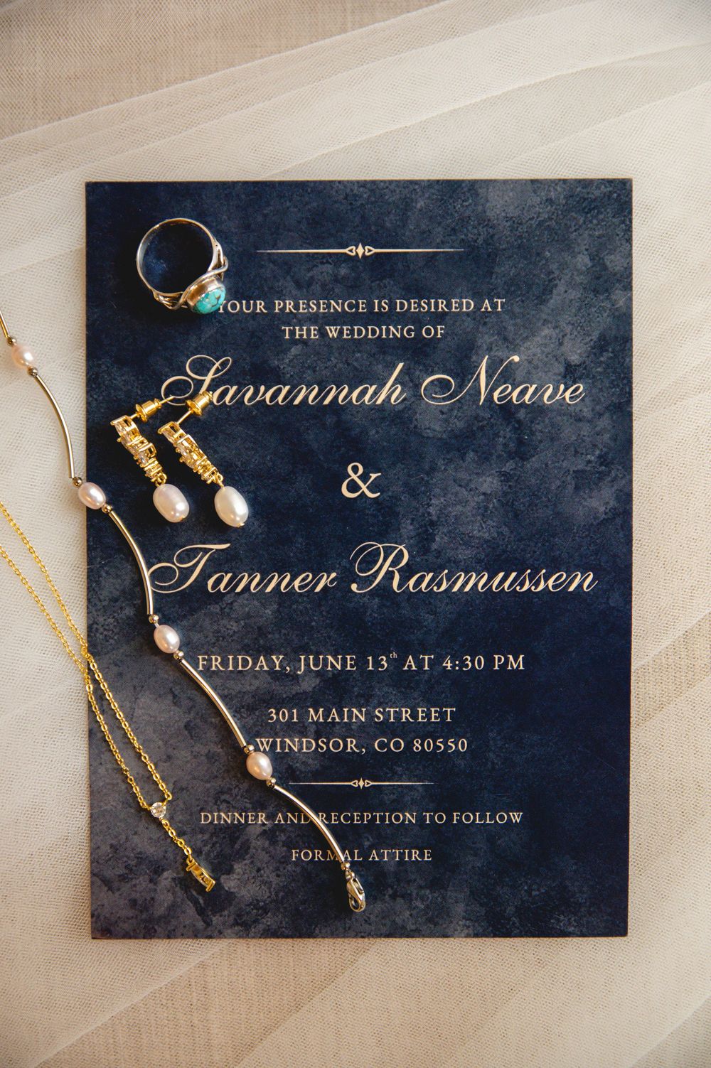 Wedding invitation details written in gold calligraphy on navy blue paper with elegant jewelry accents.