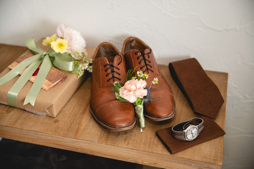 Brown leather dress shoes with floral boutonniere next to wrapped gifts and accessories on wooden surface.