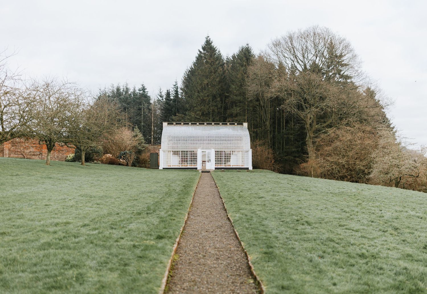 A white minimalist cottage sits at the end of a graveled path through a green lawn, surrounded by autumn trees.