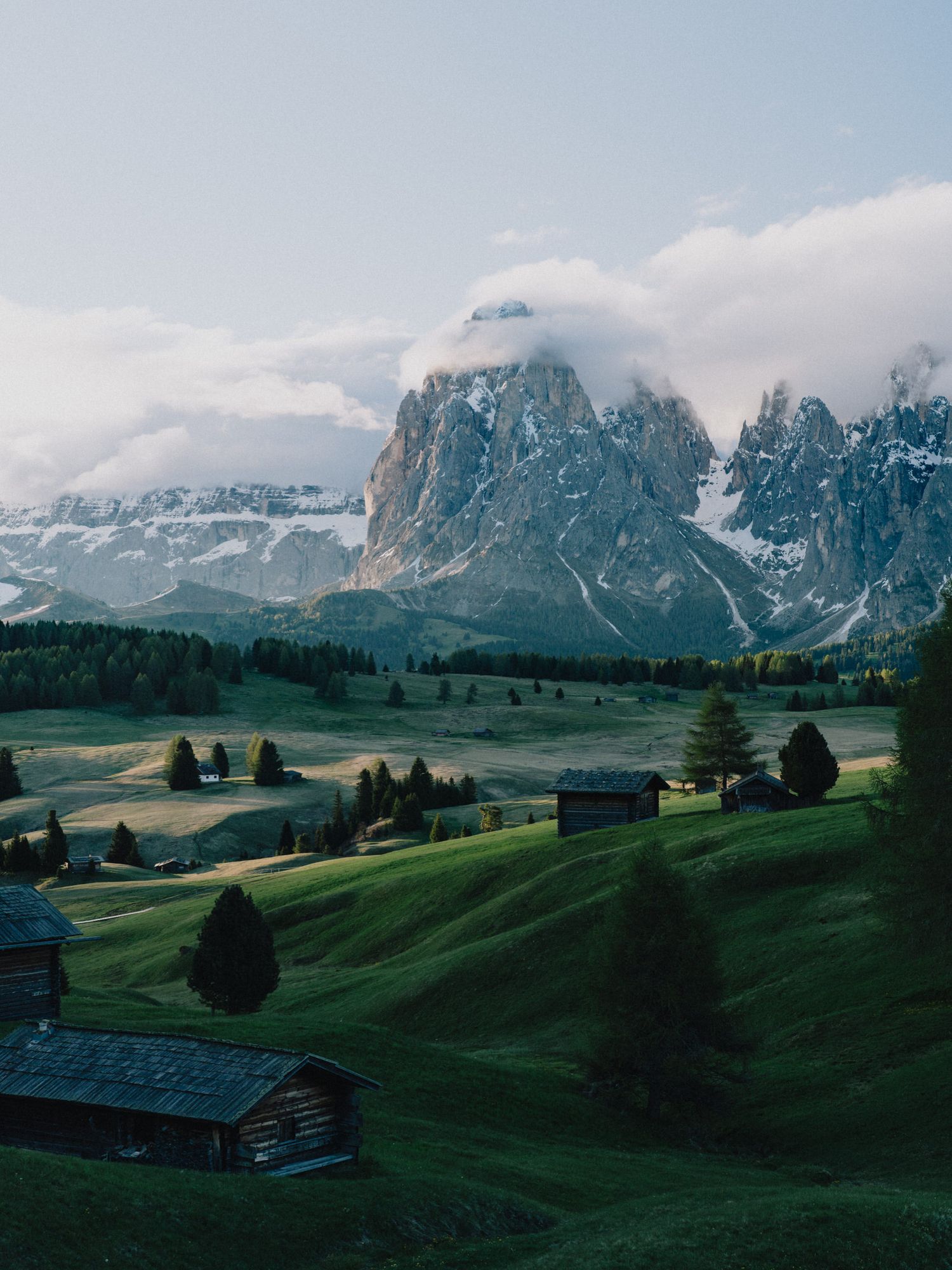 Dramatic mountain peaks rise above green alpine meadows with traditional wooden cabins in the Dolomites, Italy.