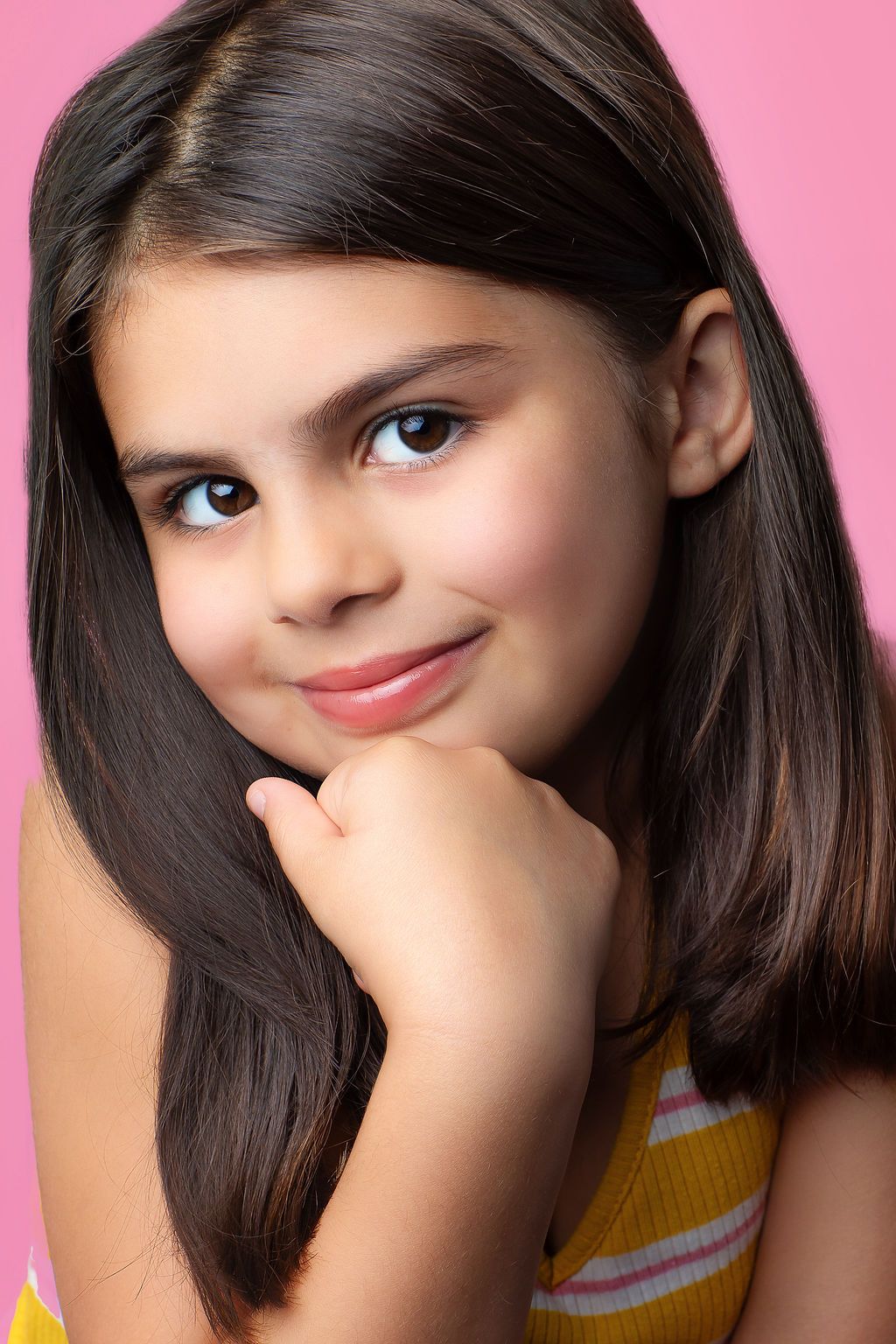 Portrait shot against pink background showing long dark hair and a warm smile with hand resting on chin.