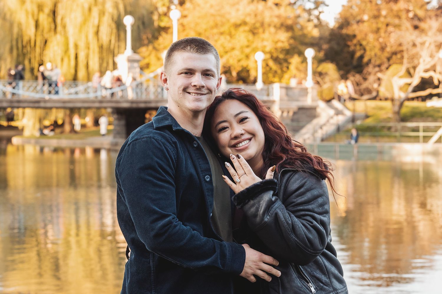 A happy couple embraces by a lake with autumn trees and ornate white bridge railings in the background.