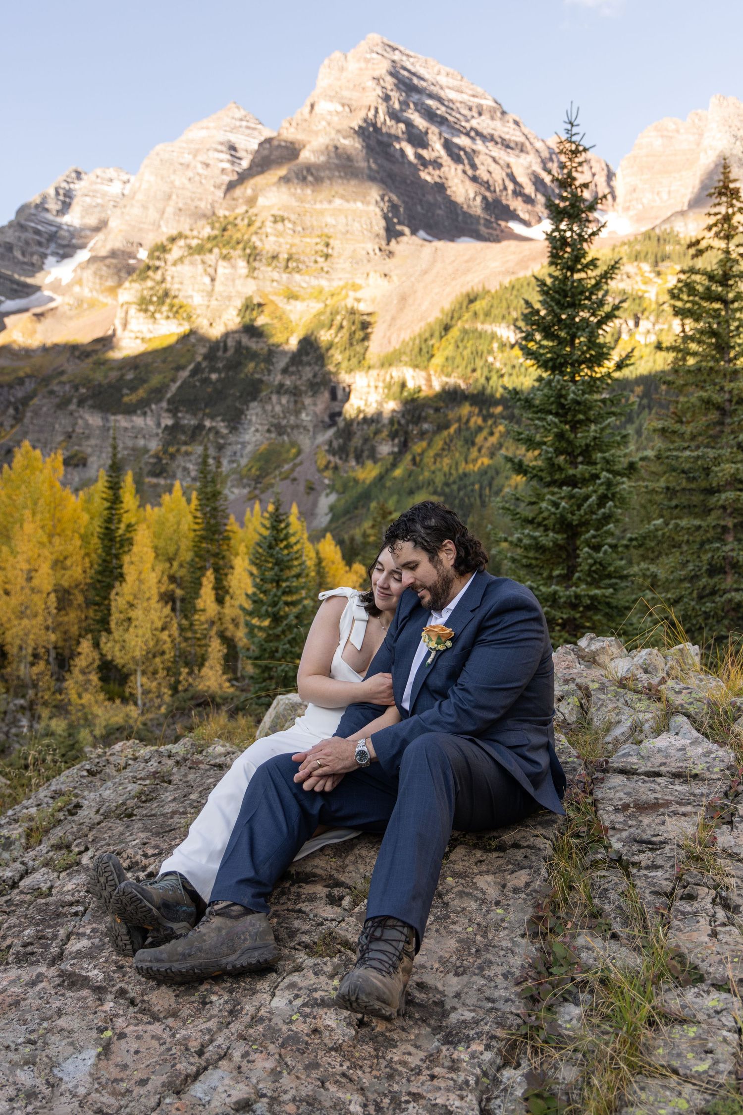 A couple shares an intimate moment while sitting on a rocky outcrop with majestic mountain peaks and autumn trees in the background.