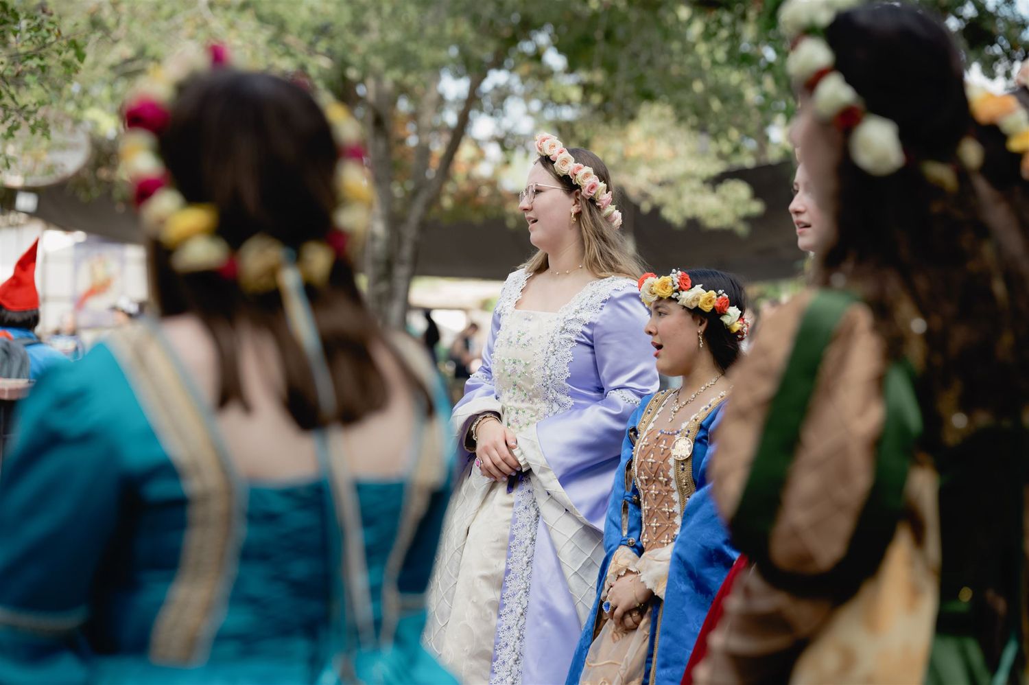 Renaissance fair participants in ornate period costumes with floral crowns gather outdoors for a festive celebration.
