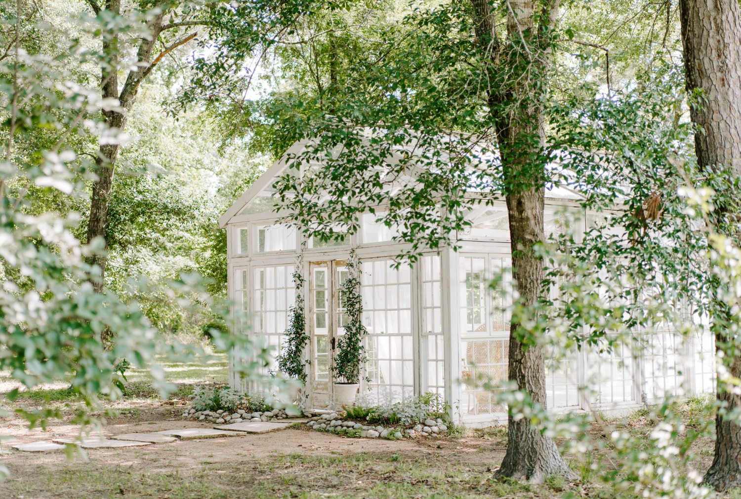 Elegant white garden greenhouse structure with arched windows nestled among lush green trees and foliage in a dreamy setting.