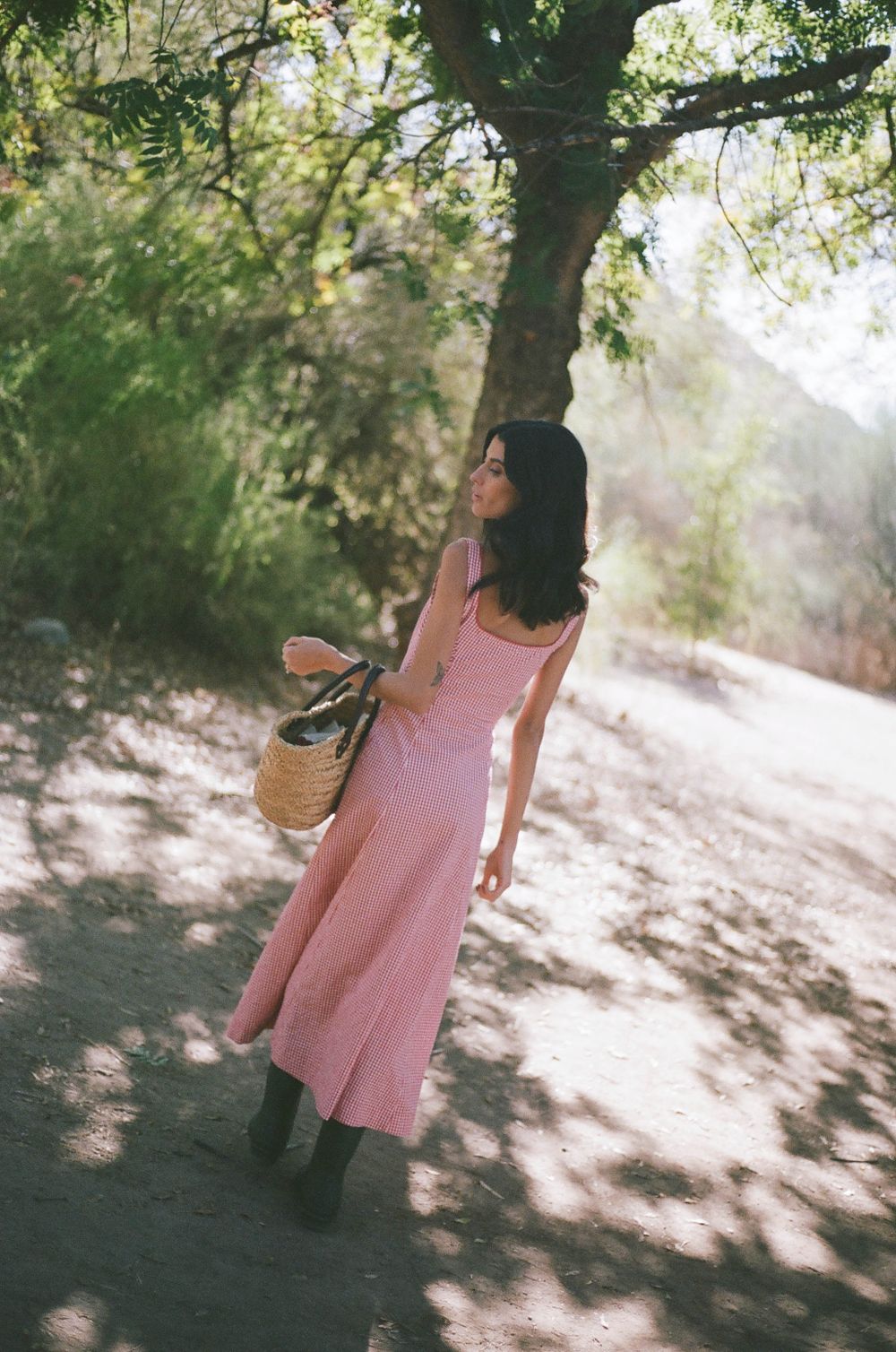 Woman in pink sundress holding wicker basket poses in dappled sunlight under trees.