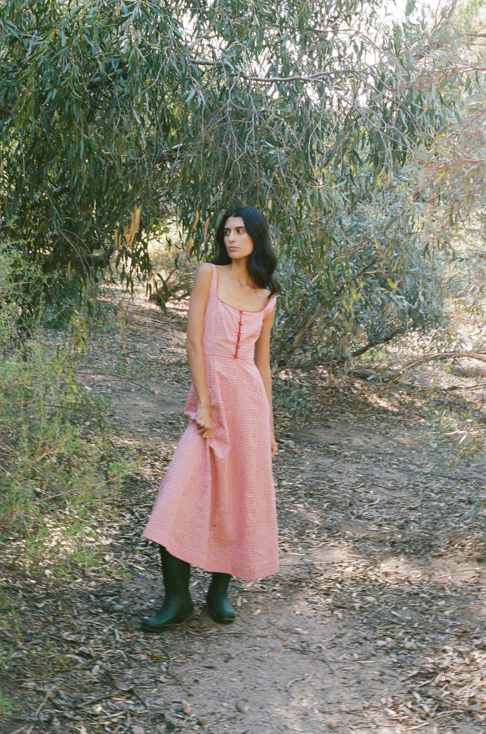 Feminine figure in pink maxi dress and green rain boots poses under willow trees in soft natural light.