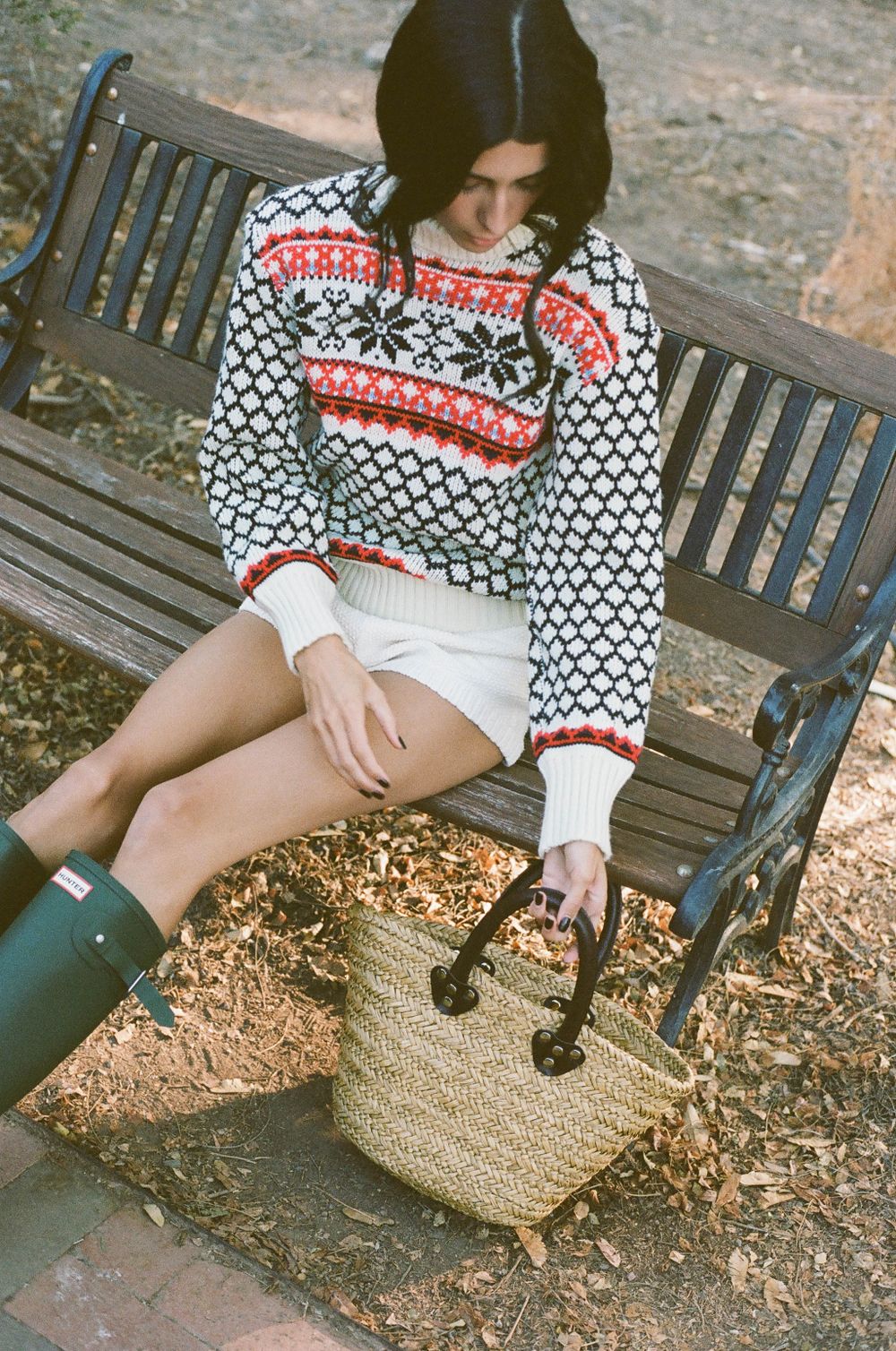 Person in festive knit sweater and shorts sitting on park bench with straw bag and rain boots.