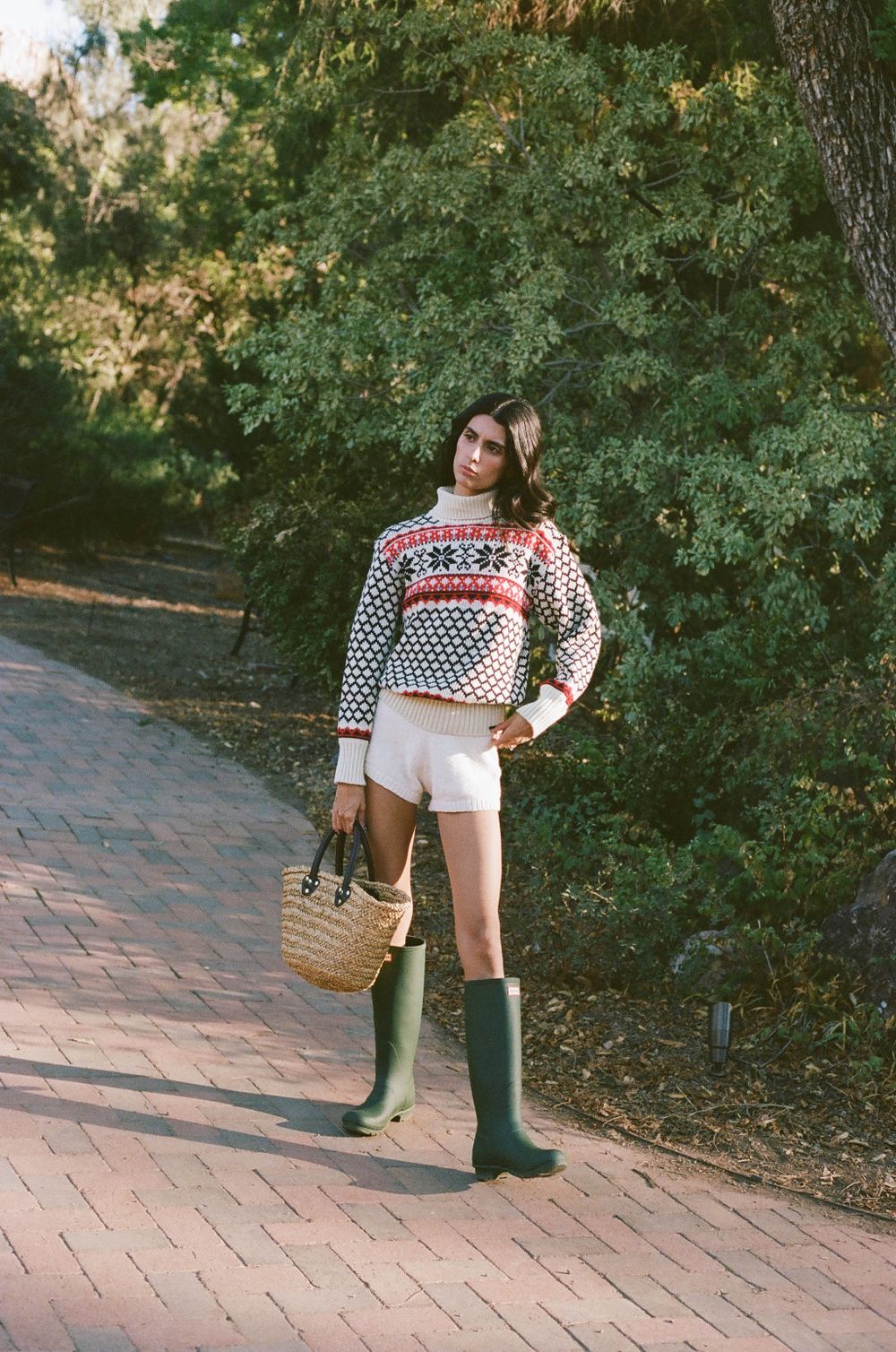 Vintage-style fashion shot with patterned sweater, white shorts and green rain boots on brick path.
