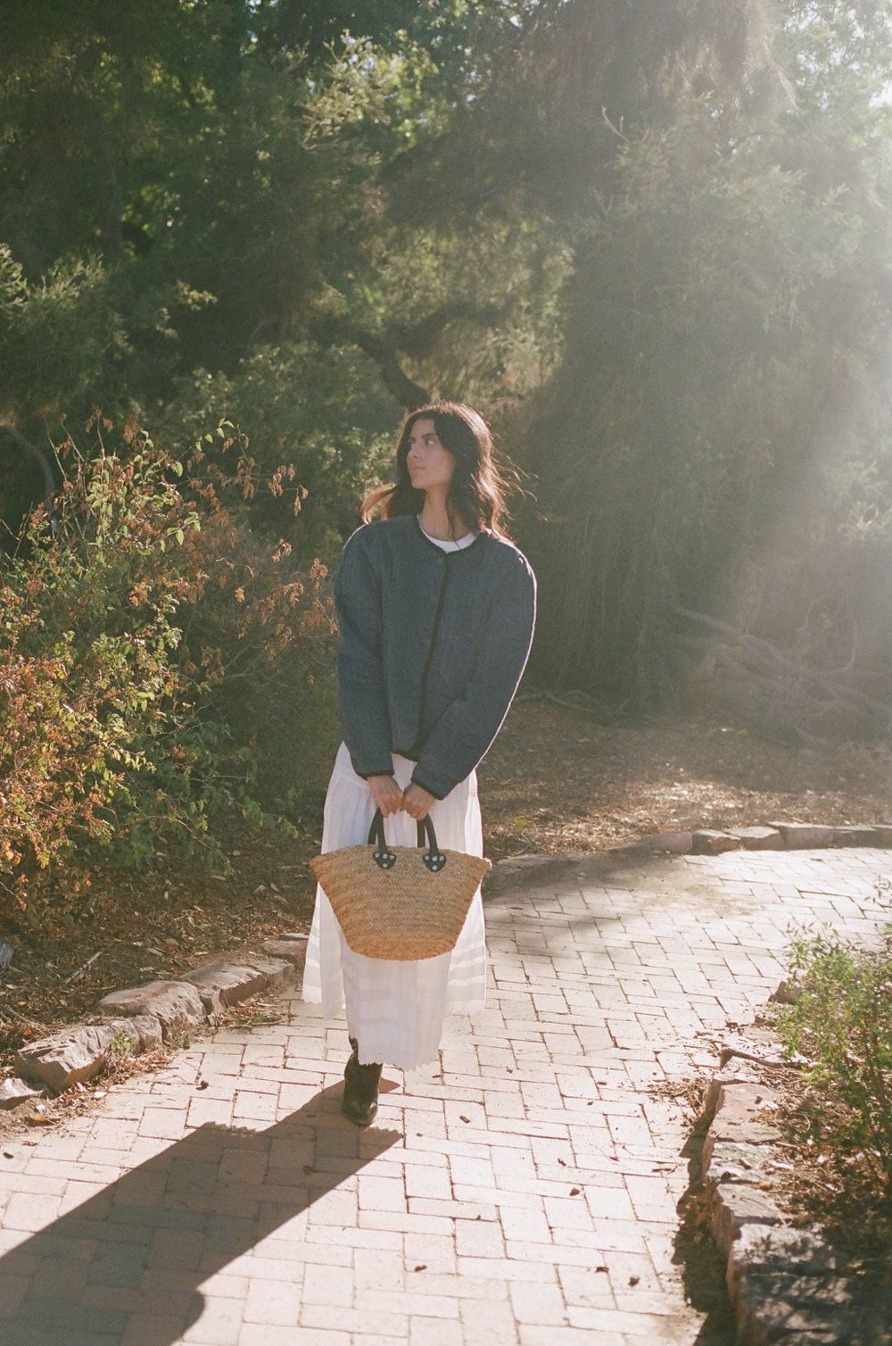 Woman in navy sweater and white dress carrying straw bag walks along sunlit brick path through trees.