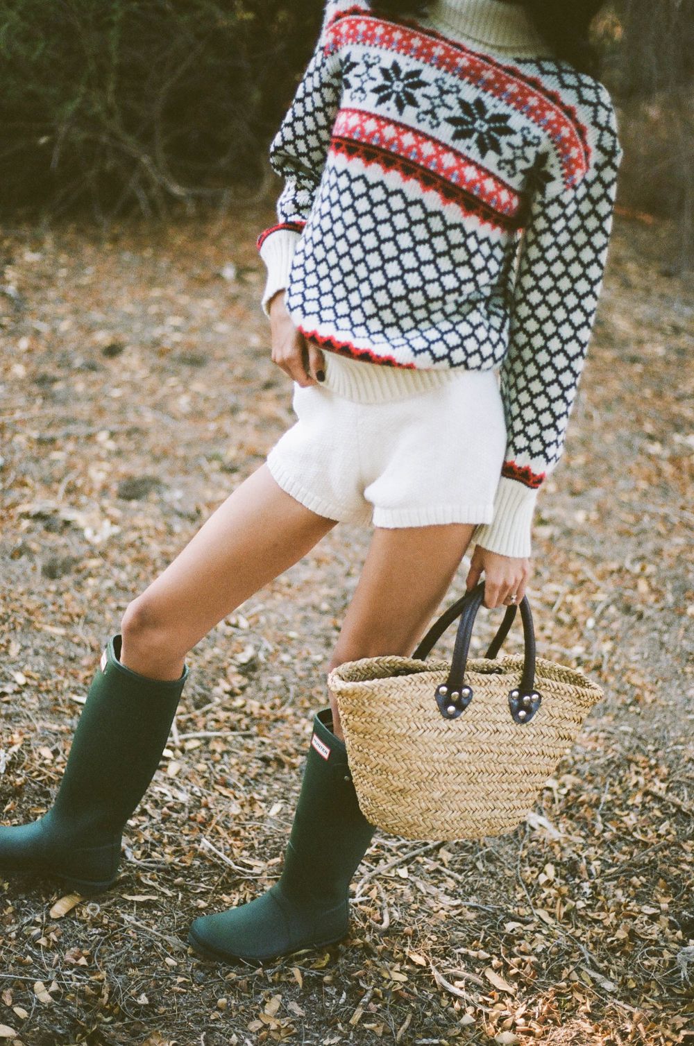 Close-up of fair isle sweater, white shorts, green rain boots and straw bag on forest path.