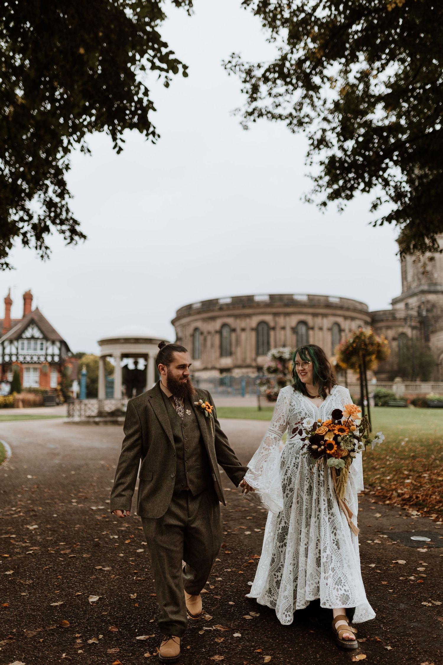 A couple walking together on an autumn day near a historic stone church, bride wearing lace dress with bouquet in The Quarry, Shrewsbury.
