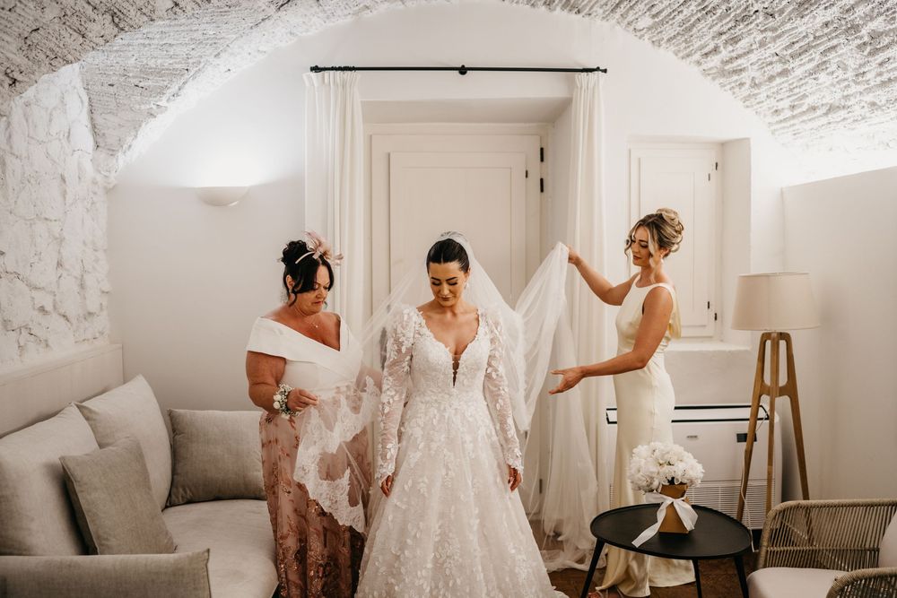 A bride in a lacy white wedding dress gets help with final preparations in an elegant white room with vaulted ceiling.