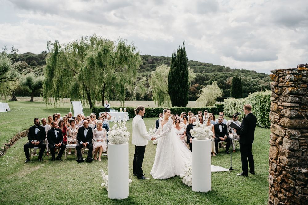 Wedding ceremony takes place outdoors on a green lawn with willow trees and rolling hills in the background.