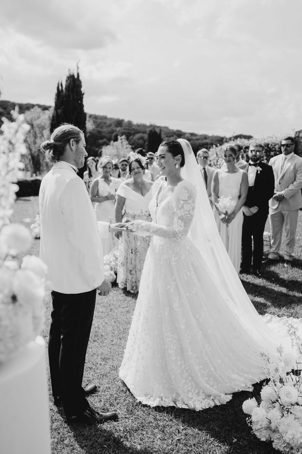 A black and white wedding ceremony photo showing an outdoor garden setting with guests watching the exchange of vows.