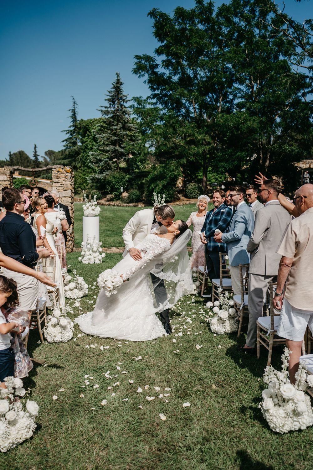 Wedding guests gather on a grassy lawn during an outdoor ceremony while flower petals scatter in the breeze.
