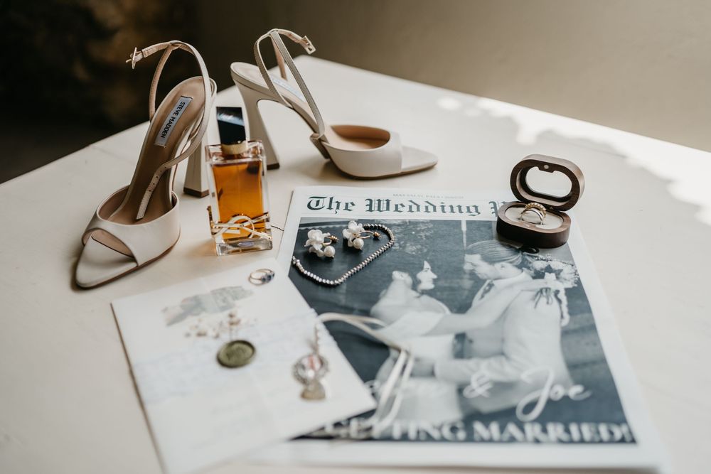 Wedding accessories including white heels, perfume, magazine and makeup compact arranged on a white surface.