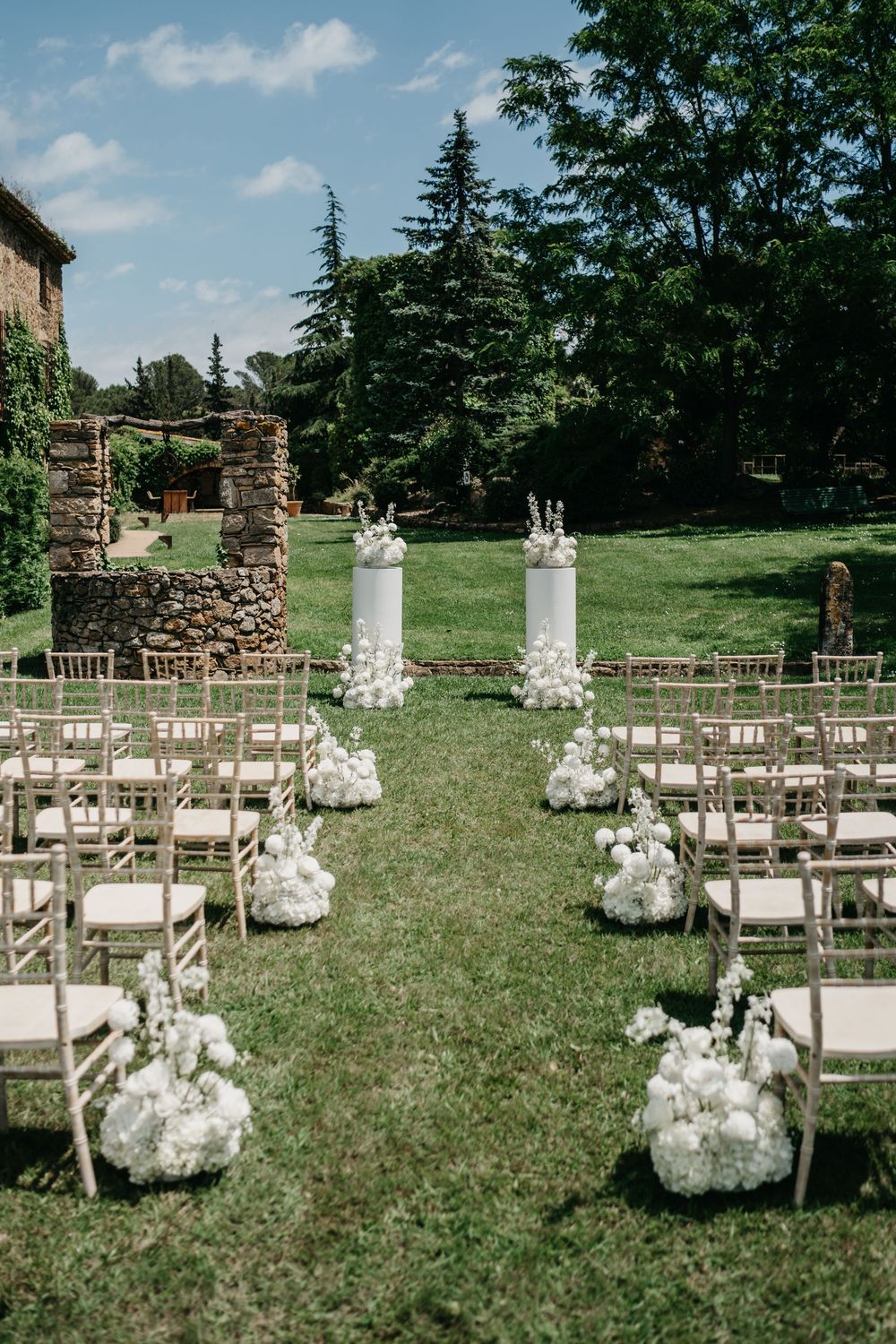 Outdoor wedding ceremony setup with wooden chairs, stone pillars and floral arrangements along the aisle on a green lawn.