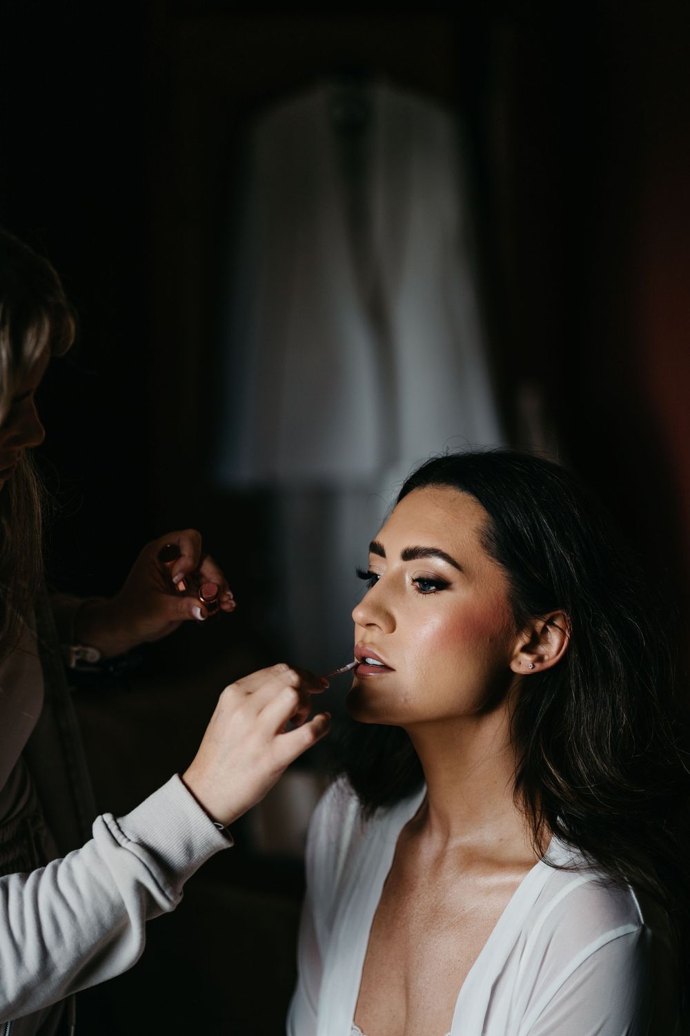 Makeup artist applies lipstick on a subject wearing a white robe in a dramatic low-light setting.
