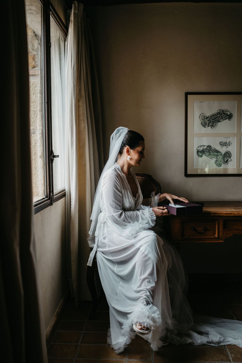 A quiet moment captured of a bride in flowing white dress seated by a window in moody lighting.