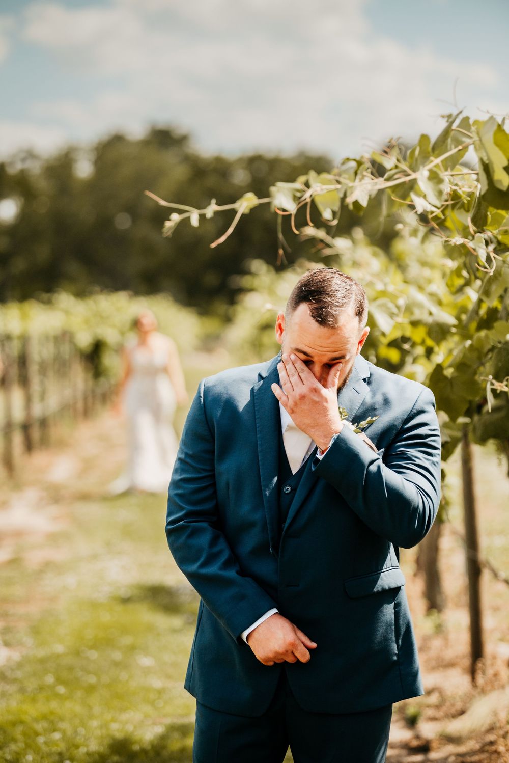 A person in a navy suit stands in a vineyard on a sunny afternoon.