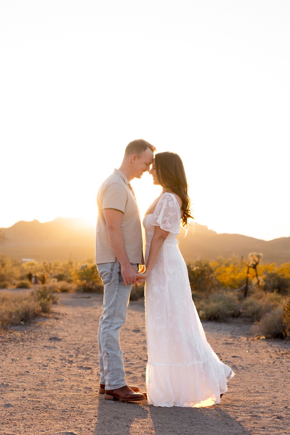 Couple in white and gray attire share romantic moments at sunset in a desert landscape while holding hands.