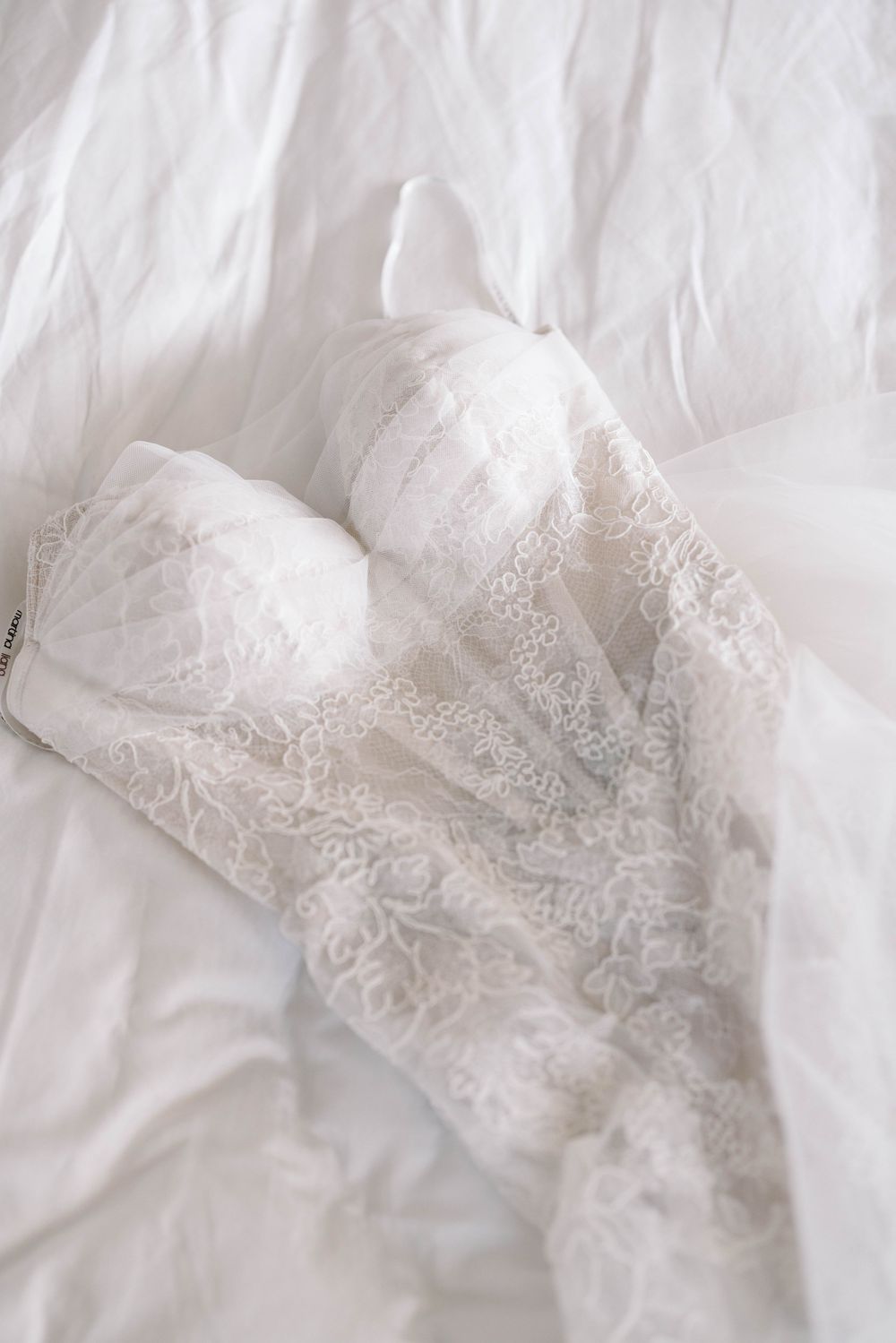 Delicate white lace bridal bustier lies on white bedding with intricate floral detailing and feminine design details.