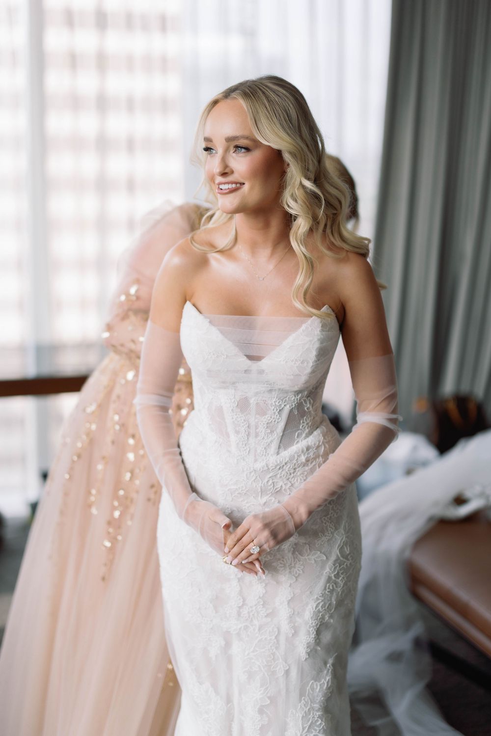 Beautiful bride in white lace strapless wedding gown smiles while getting ready in bridal suite.