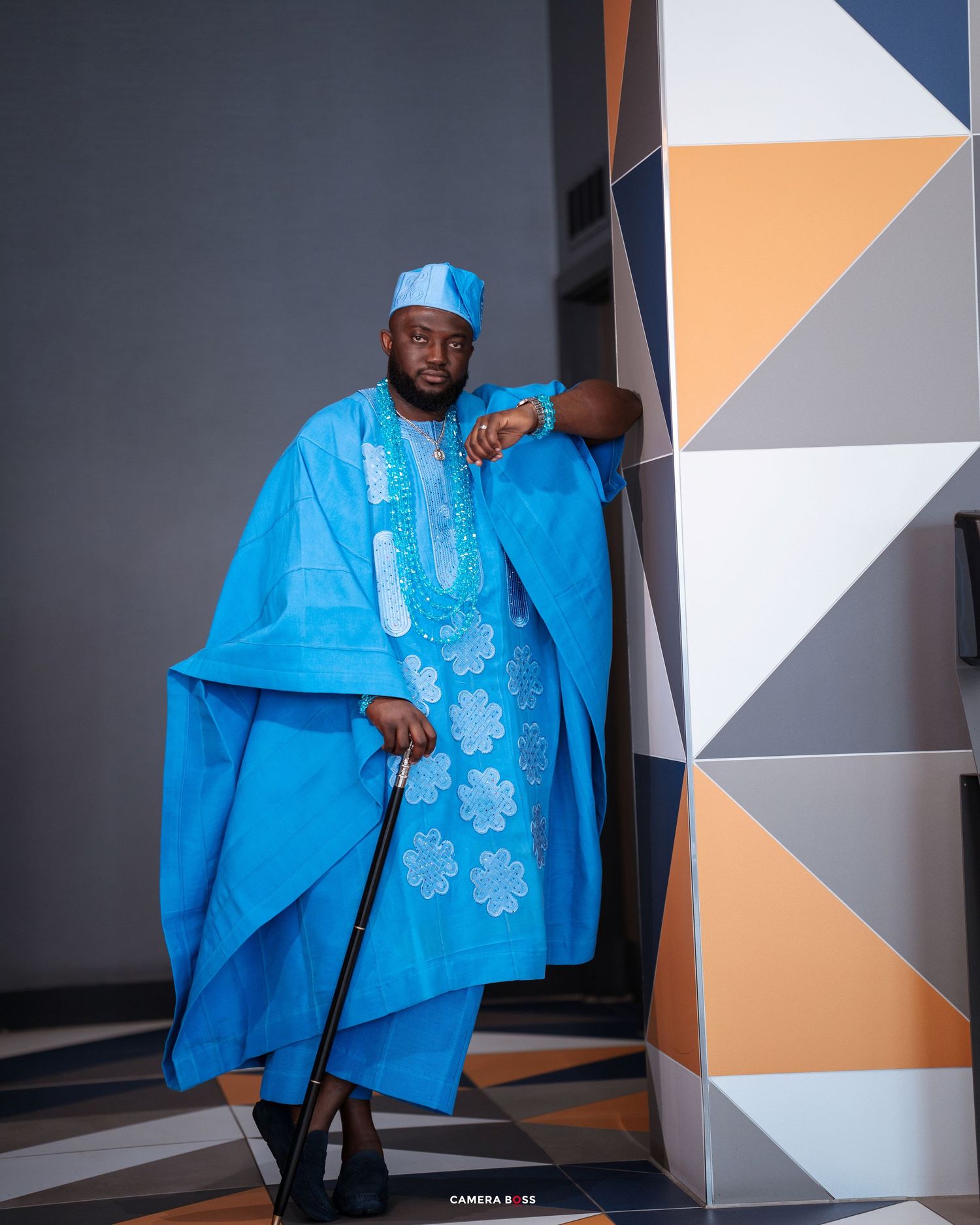 Traditional African male attire featuring a bright blue agbada with embroidered details and matching cap against geometric wall art.