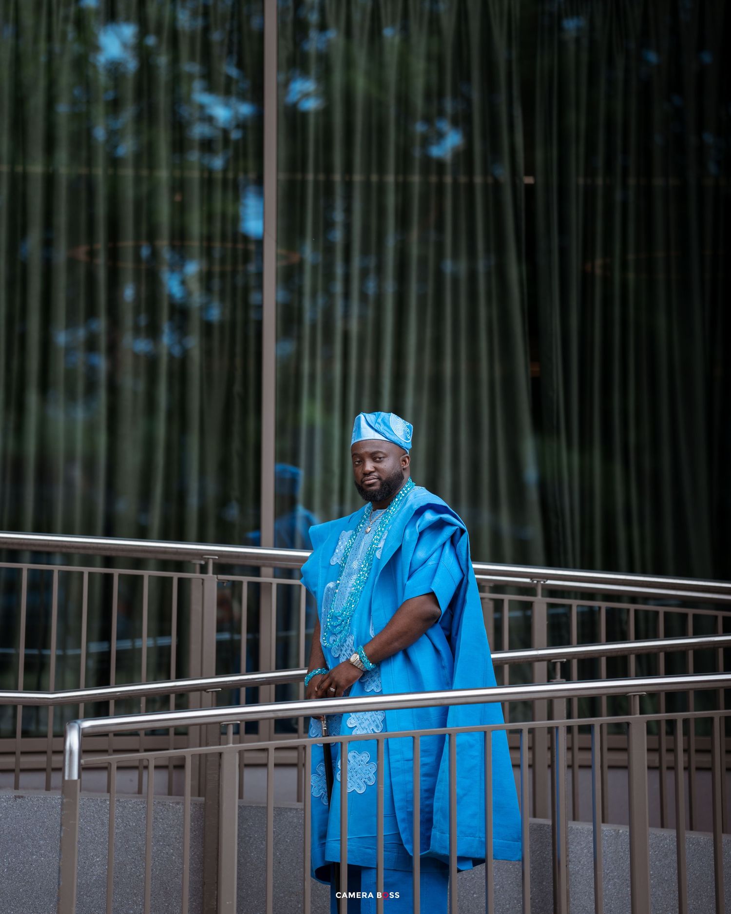 Person in vibrant blue traditional African agbada attire stands against bamboo forest background.