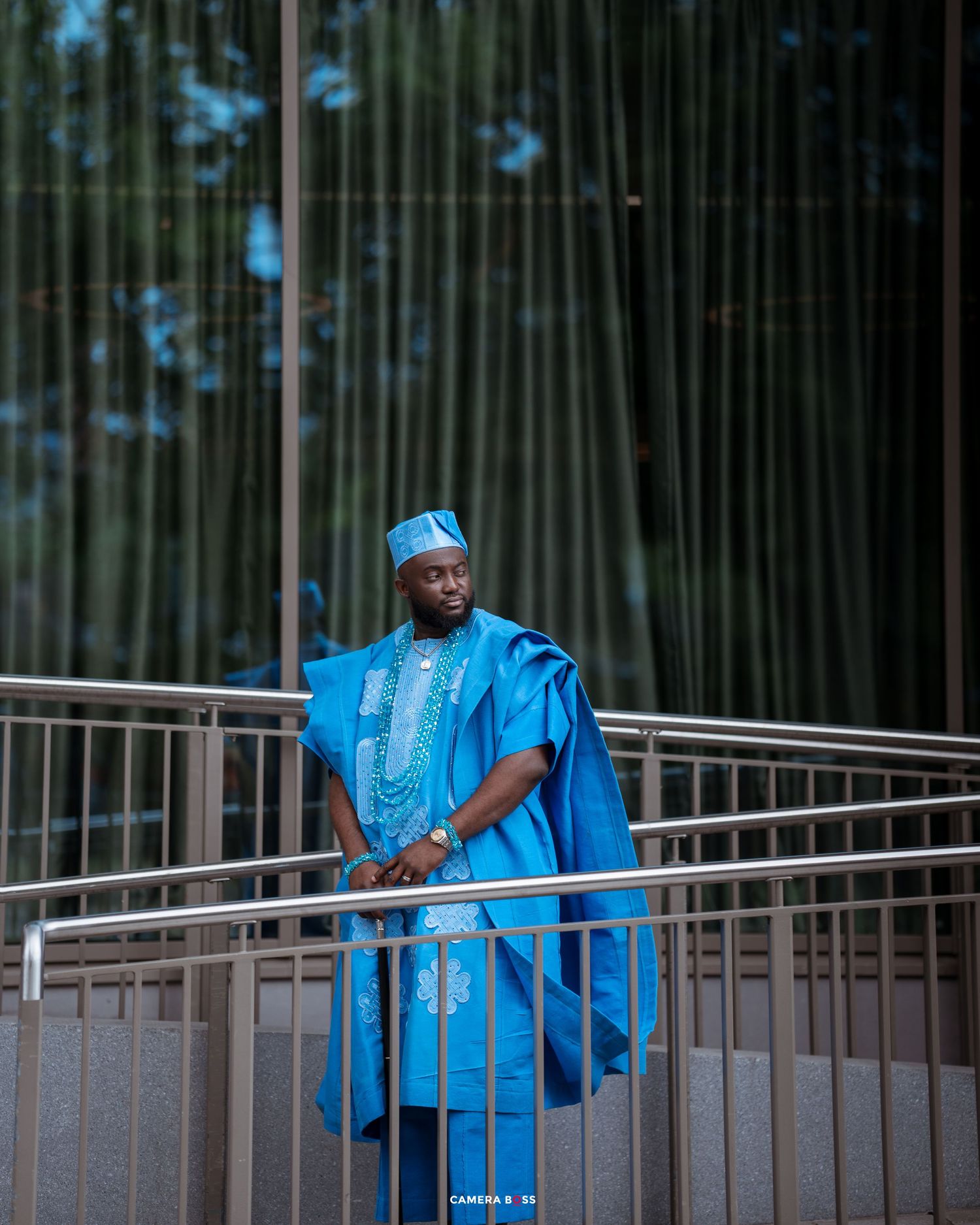 Person wearing vibrant blue traditional African agbada outfit stands confidently on a balcony with bamboo backdrop.