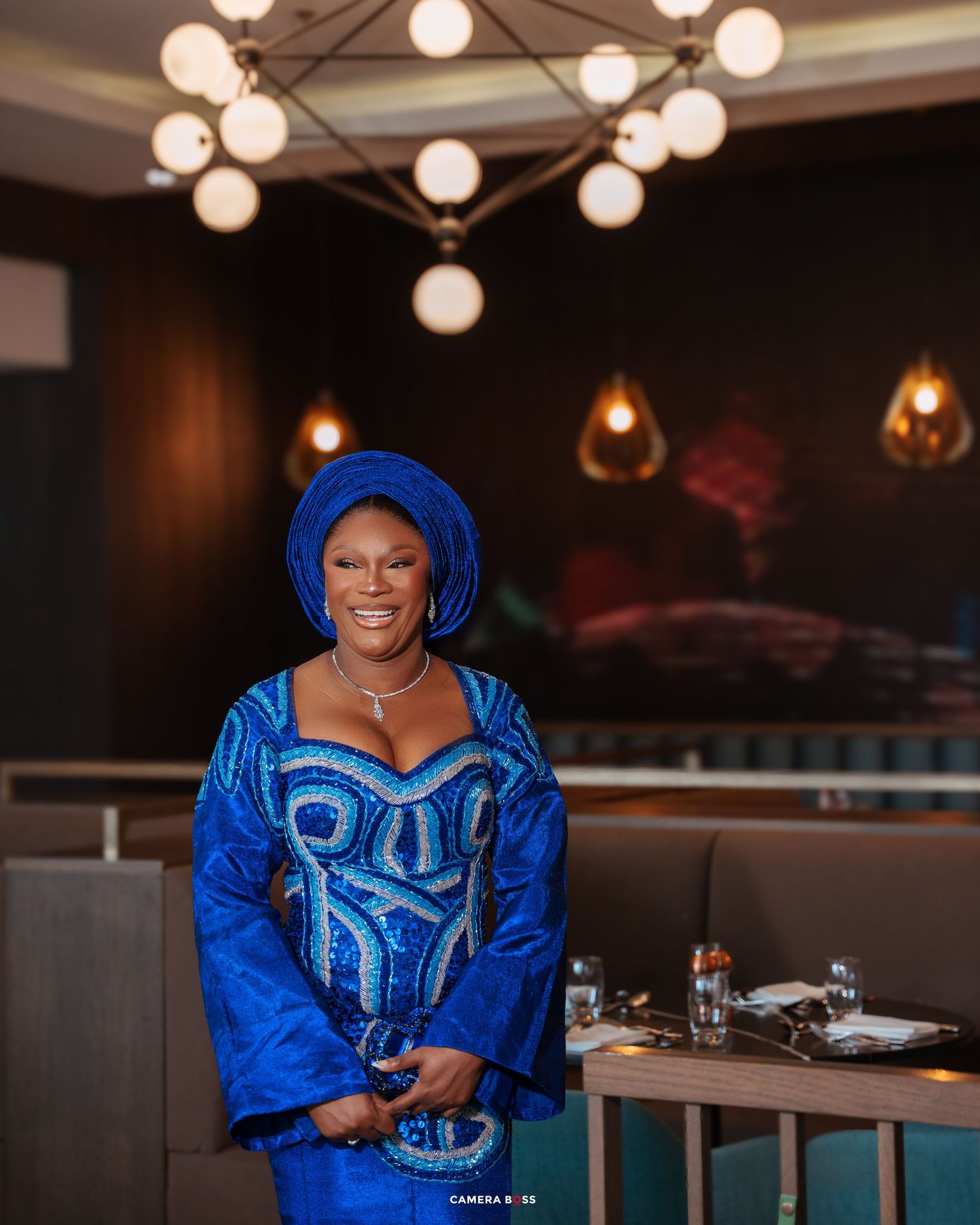 Person in vibrant royal blue traditional African attire with embroidered details poses inside a restaurant with modern lighting.
