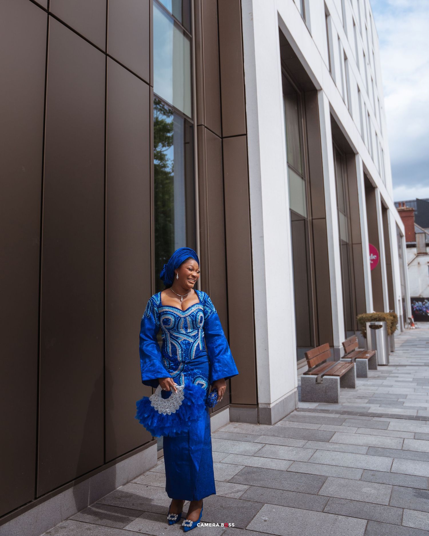 Person wearing a blue traditional African dress and headwrap poses elegantly outside a modern building.