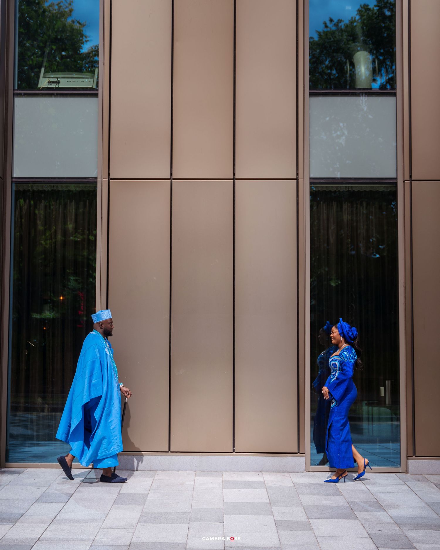 Two people in vibrant blue traditional garments walk past a modern building exterior.