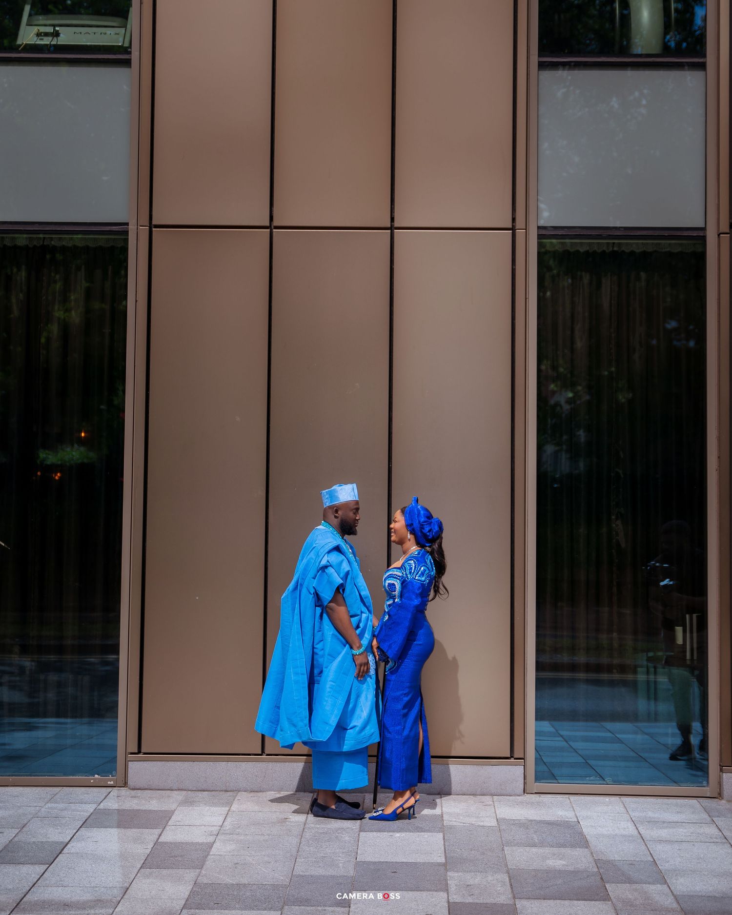 Two figures in vibrant blue traditional African clothing walk along a modern building's exterior walkway.