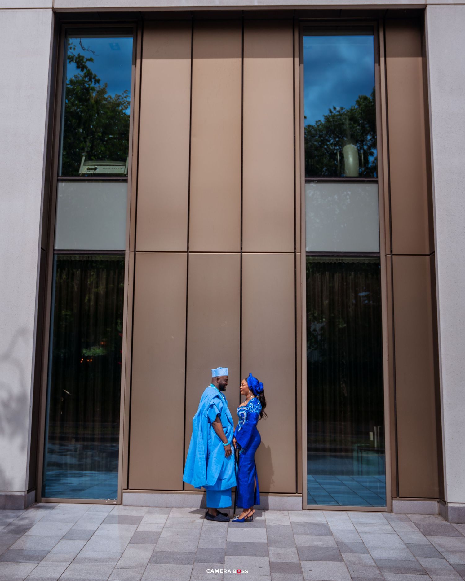 Couple in matching blue traditional African attire embrace in front of a modern building with reflective windows.