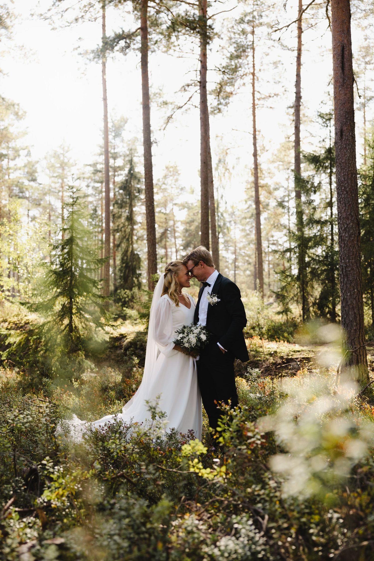 A couple stands together in a sunlit forest clearing surrounded by tall pine trees.