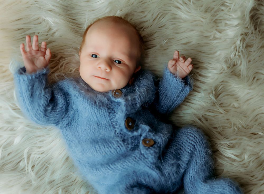 A baby in a soft blue knit romper lies on a fluffy white fur blanket with arms outstretched.