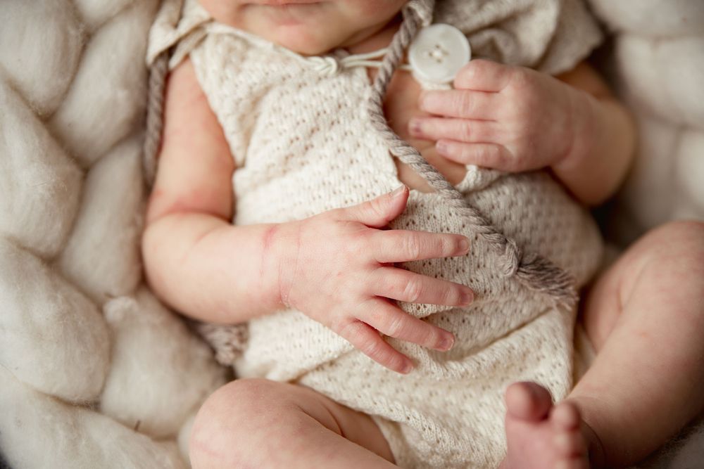 Close-up of tiny hands grasping cream-colored knitted fabric against soft white textured background.