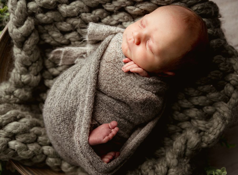 Sleeping newborn wrapped snugly in gray swaddle blanket on chunky knit charcoal throw blanket.