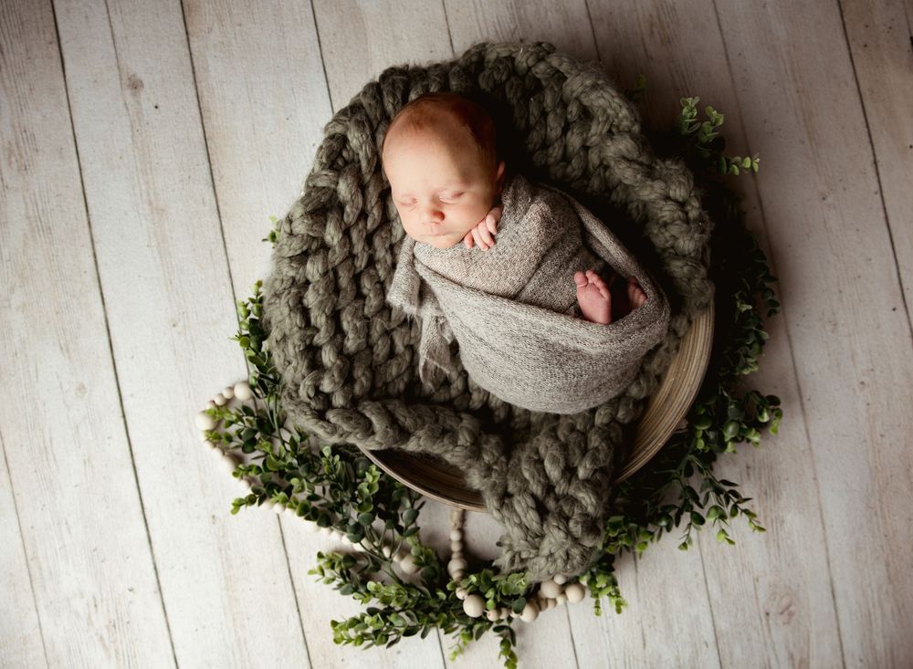 Newborn baby peacefully sleeping in gray wrap on wooden bowl surrounded by chunky knit blanket and greenery wreath.