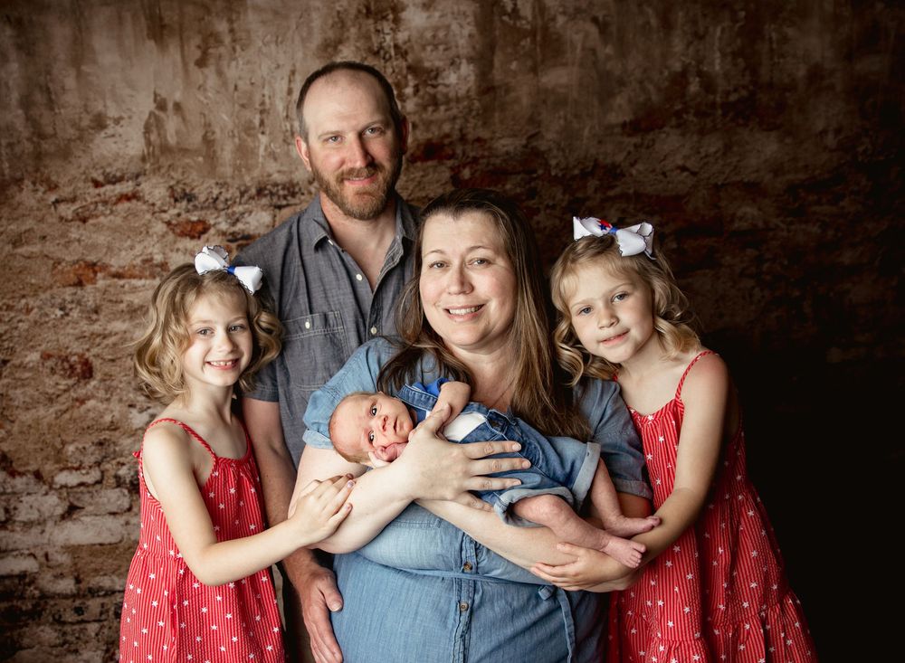Family portrait with newborn baby features matching red dresses on two young girls against rustic brick wall background.