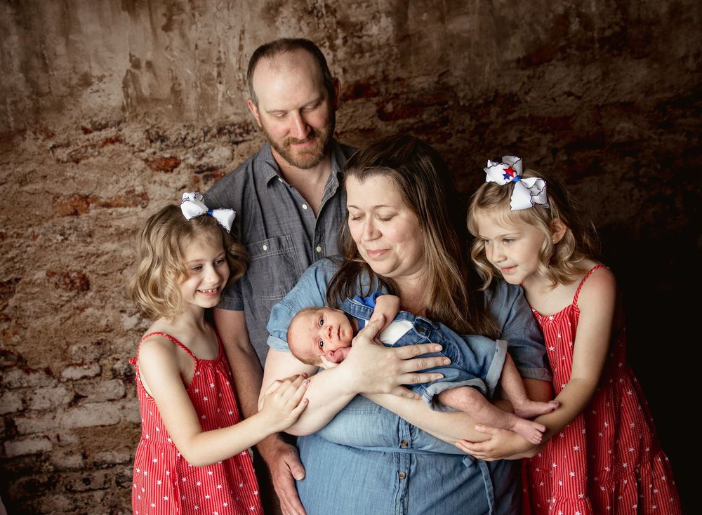 Family gathered in rustic setting with brick wall background and three young children wearing red dresses.