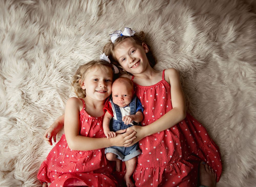Baby in red lace dress and navy blue bloomers lies on soft cream fur rug while laughing and playing.