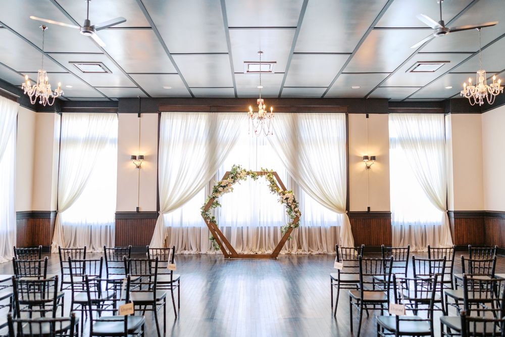 An elegant wedding venue with a hexagonal floral arch, rows of black chairs, and white drapes against a dark wood floor.