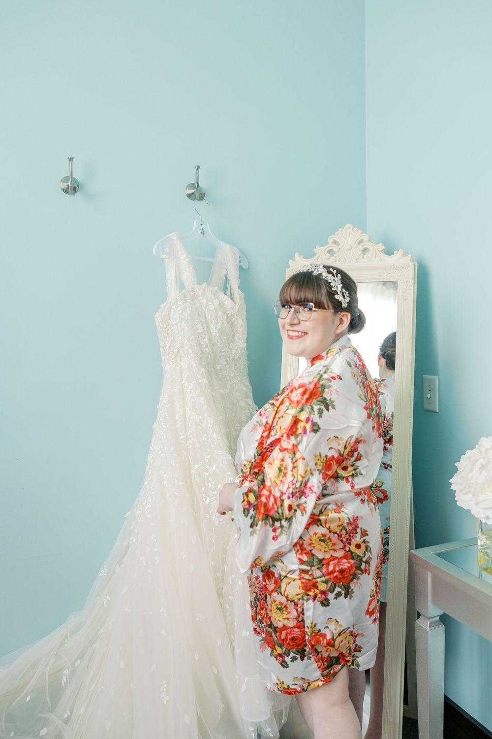 A wedding gown hanging next to an ornate mirror reflects a floral robe against light blue walls in a bridal prep room.