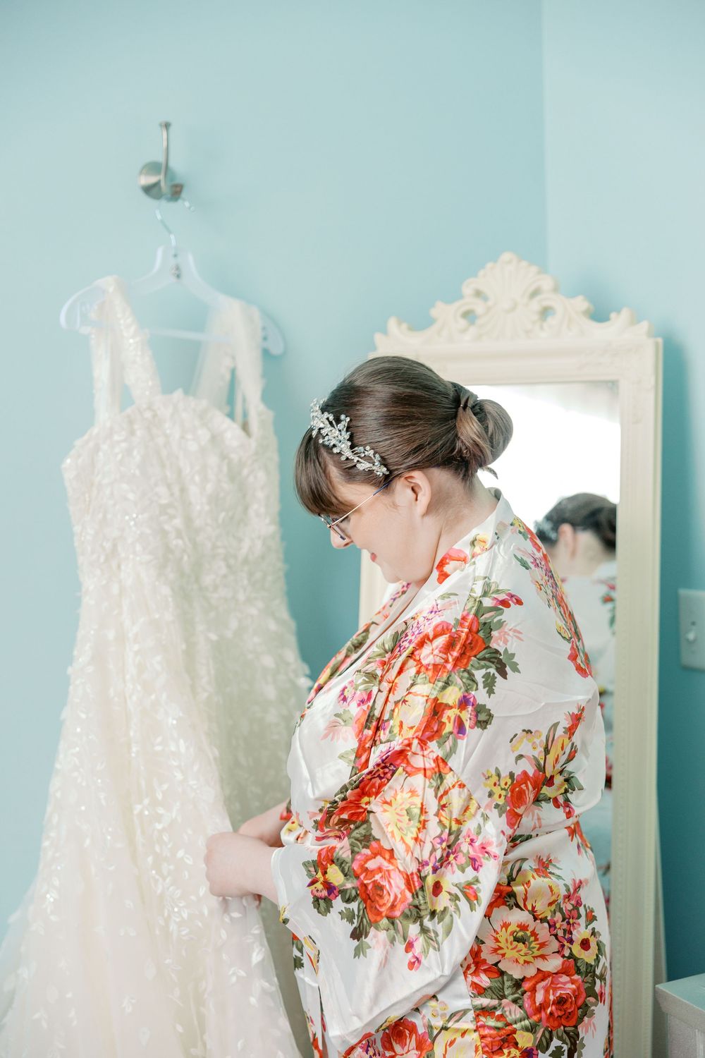 Lace white wedding dress hangs beside mirror while someone in floral robe admires it in a blue room.